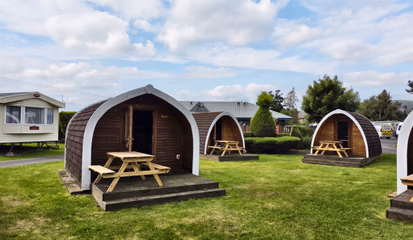 Three timber camping pods in a row on mown grass, each with its own picnic bench